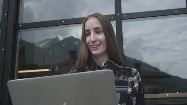 A young beautiful girl uses a laptop for a video call in a cafe against the backdrop of mountains. Business call or video meeting with parents. Downshifting during a pandemic and quarantine.