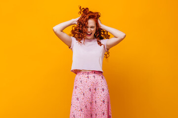 Perky girl in pink T-shirt and skirt ruffles her red hair against orange background