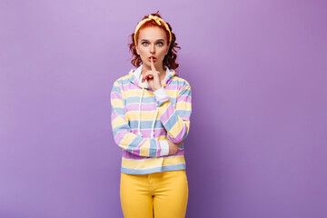 Blue-eyed woman in yellow pants and multi-colored sweatshirt puts her finger to her mouth asking to observe silence