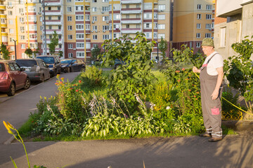 A gardener in overalls with a yellow hose watering flowers against the background of apartment...