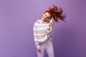 Beautiful girl in skinny jeans and sweatshirt holding apple and playing hair against lilac background