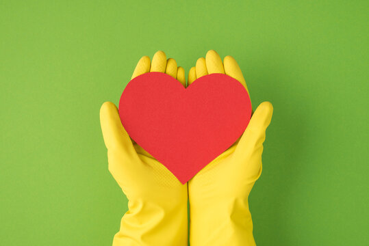 Overhead photo of hands in yellow gloves holding the pink card as heart isolated on the green background - Powered by Adobe