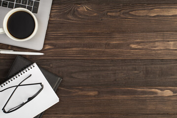 Top view photo of cup of coffee on laptop white pen glasses on two black and white notepads on isolated dark wooden table background with copyspace