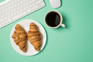 Overhead photo of keyboard computer mouse cup of coffee and plate with two croissants isolated on the teal backdrop