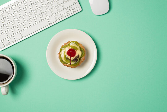 Overhead photo of keyboard computer mouse cup of coffee and plate with fruit cake isolated on the teal backdrop