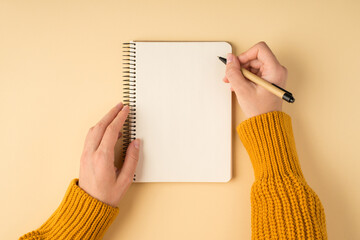 First person top view photo of female hands in yellow pullover writing in spiral notepad on isolated light orange background with copyspace