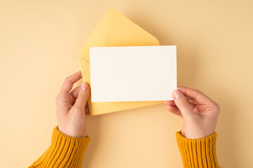 First person top view photo of woman's hands in yellow pullover holding open pastel yellow envelope and white card on isolated light orange background with blank space