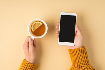 First person top view photo of female hands in yellow sweater holding white cup of tea with lemon...