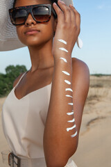 dark-skinned woman applying white cream on her hand while standing on the sand in the summer