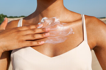 dark-skinned woman applying white cream to her chest while standing on the sand in the summer