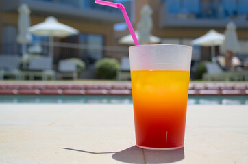 A bright orange cocktail with a pink straw stands by the pool.