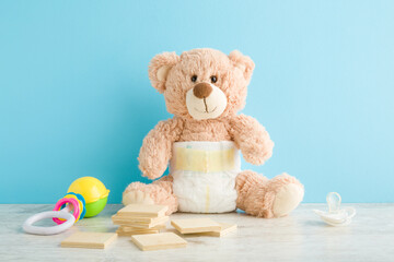 Rattle toy, playing wooden shapes, soother and brown teddy bear in white baby diaper on table at light blue wall background. Pastel color. Closeup. Front view.