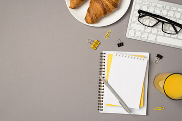 Top view photo of workstation glass of juice plate with croissants glasses on keyboard stationery binder clips and pen on stack of organizers on isolated grey background with blank space - Powered by Adobe