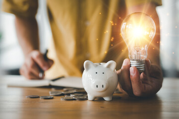 saving energy and money concept. idea for save or investment. businessman holding lightbulb beside piggy bank and coins stacking on desk with note book. - obrazy, fototapety, plakaty