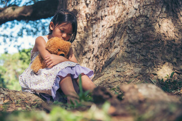 Sad girl hugging teddy bear sitting under tree sadness alone in green park. Lonely girl feeling sad...