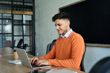 Young Indian latin happy smiling student programmer developer at desk working using pc laptop computer studying online class at modern office university campus. Remote education, virtual learning.