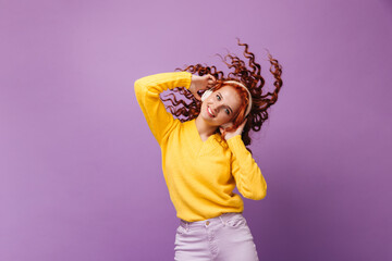 Charming woman in yellow sweater smiles, listens to songs in headphones and dances against lilac background