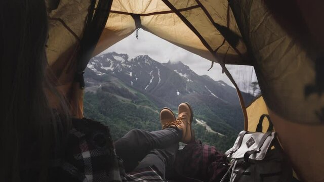 Female legs of a hiker in yellow boots lie in a yellow tent. The traveler's legs are resting, the girl lies and admires the mountains after the ascent. Feet and big mountains. Rest in the camp.