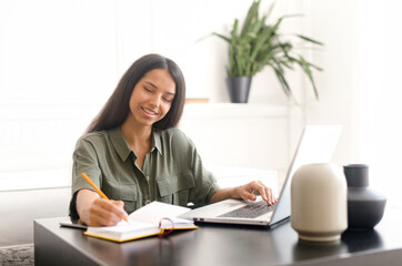 Smiling indian female student using laptop for watching webinars, lectures, sits at desk in home...