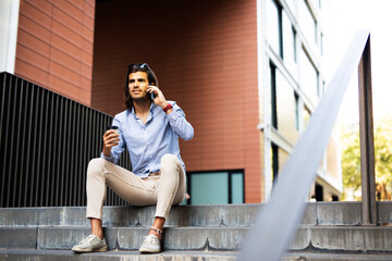 Young man sitting on the stairs drinking coffee. Handsome businessman using the phone while drinking coffee..