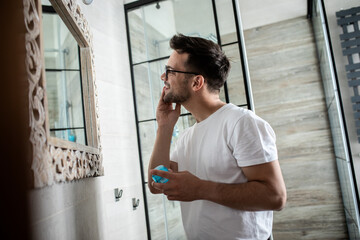 Young man going through morning routine in the bathroom applying face cream.