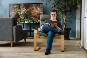 Young man with down syndrome reading book while sitting in armchair