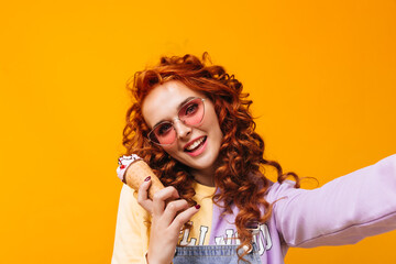 Cheerful curly girl in sweatshirt and pink glasses smiles, takes selfie and holds ice cream