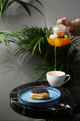 eclair on ceramic plate anf cup of tea on marble stone and tropic plants on background. tea pot pouring. woman hand hold teapot. tea splash
