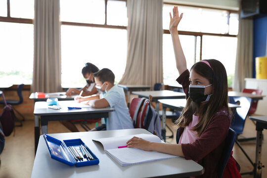 Caucasian girl wearing face mask raising her hand while sitting on her desk in the class at school