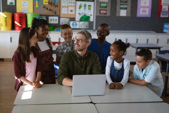 Caucasian male teacher and group of diverse students smiling while using laptop in class at school