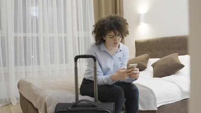 Young woman sitting on bed in hotel room, scrolling smartphone, travel, taxi app