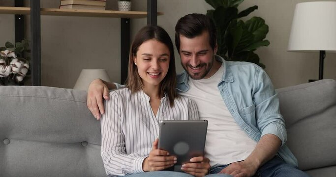 Affectionate loving millennial gen family couple resting on cozy sofa, looking at tablet screen, involved in pleasant video call web camera zoom conversation, modern tech distant communication concept
