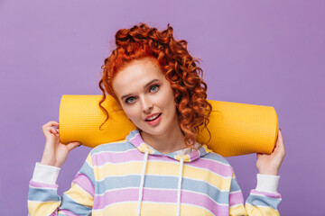 Blue-eyed woman in striped sweatshirt holds yellow sport mat and looks into camera