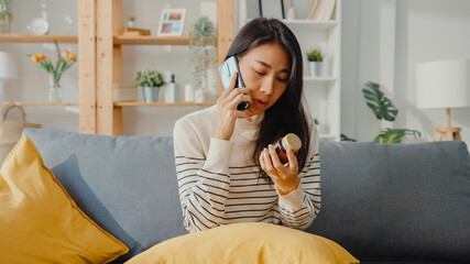 Sick young Asian woman hold medicine sit on couch use smartphone call to consult with doctor at...