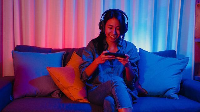 Happy Asia Girl Gamer Wear Headphone Competition Video Game Online With Smartphone Excited Talk With Friend Sit On Couch In Colorful Neon Lights Living Room At Home, Home Quarantine Activity Concept.