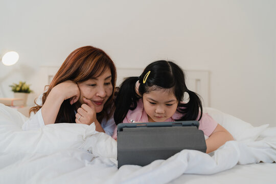 Asian grandmother using tablet read fairy tales to granddaughter at home. Senior Chinese, grandma happy relax with young girl before bedtime lying on bed in bedroom at home at night concept.