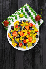 Fruit salad in a white plate, bowl on a napkin made of fresh fruits from grapes, nectarine, kiwi and strawberries, blueberries, honeysuckle on a dark wooden background.Top view, copy space