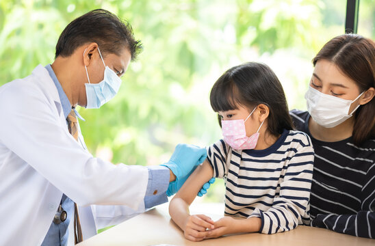 Asian  Senior Doctor Wearing Gloves And Isolation Mask Is Making A COVID-19 Vaccination In The Shoulder Of Child Patient With Her Mother At Hospital.