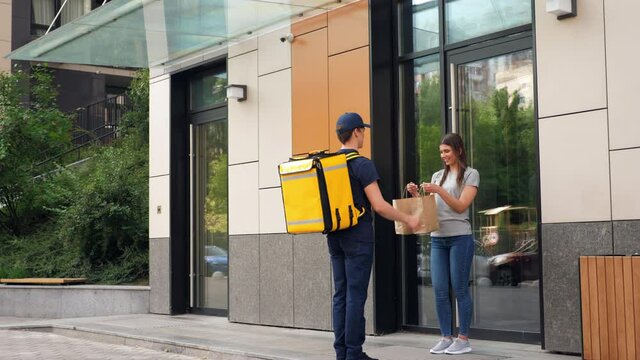 Wide shot positive food delivery man courier with yellow thermal backpack deliver order parcel to happy woman customer. Deliveryman worker delivers lunch to smiling client. Online internet shopping