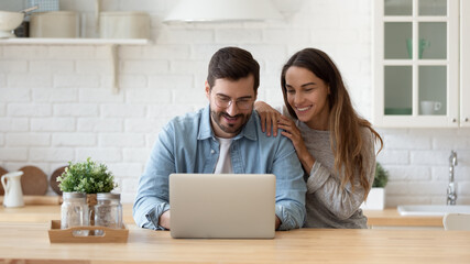 Attractive 35s couple sit at table in cozy kitchen looking at laptop screen, buying goods, tickets online, using e-commerce retail e-services feel satisfied, enjoy comfort usage of modern technology