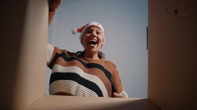 Young person with santa hat looking inside package getting excited about gift and christmas season celebration. Caucasian woman opening box with present from relatives being happy. View from inside