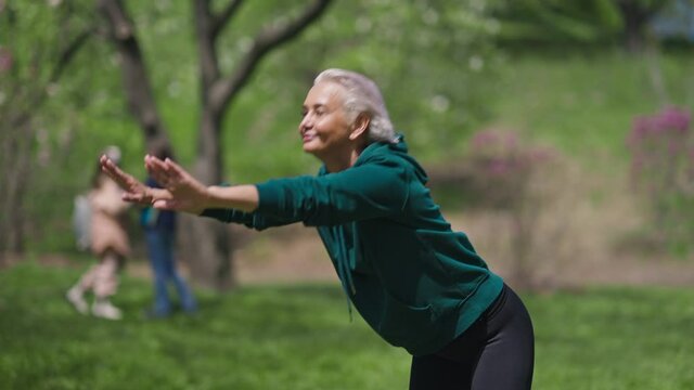 Positive slim female retiree bending forward smiling working out in sunny summer spring park. Portrait of happy joyful Caucasian woman training exercising outdoors. Sport and retirement