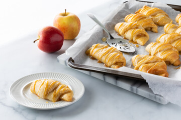 Apple filled crescent roll on a small plate with a baking sheet of more in behind, against a bright...