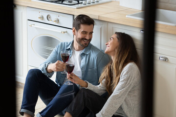 Beautiful young couple in love celebrate relocation day to own or rented home resting sit on warm floor in kitchen clinking glasses of red wine feel satisfied glad to start new life at modern house