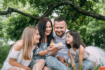 Caucasian father, mother and two little daughters eating homemade cupcakes during picnic at green garden. Happy family enjoying sweet dessert on fresh air.