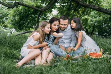 Fototapeta premium Caucasian father, mother and two little daughters eating homemade cupcakes during picnic at green garden. Happy family enjoying sweet dessert on fresh air.