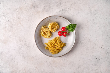 Uncooked raw girandole, tagliatelle, cherry tomatoes and basil on white plate on grey textured background. Copy space. Top view