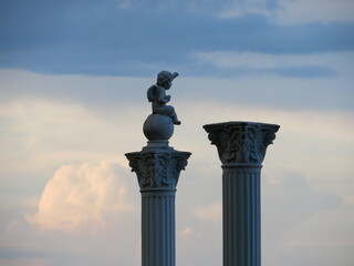 an angel sits on a ball on a Roman column, two columns against a background of blue sky and clouds