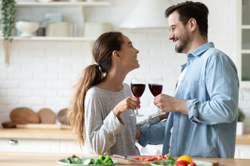 Beautiful millennial couple in love holding glasses of red wine celebrate special occasion standing in modern kitchen enjoy romantic date feel happy and enamoured. House-warming, anniversary concept
