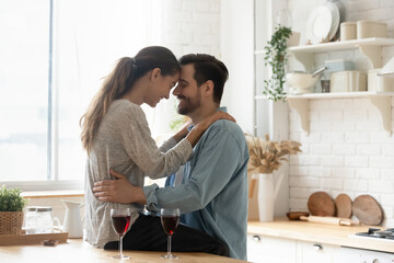Lovely married couple in love touch foreheads while spend time together at modern home enjoy romantic dating in kitchen with red wine. Romance, anniversary celebration, life event, relations concept